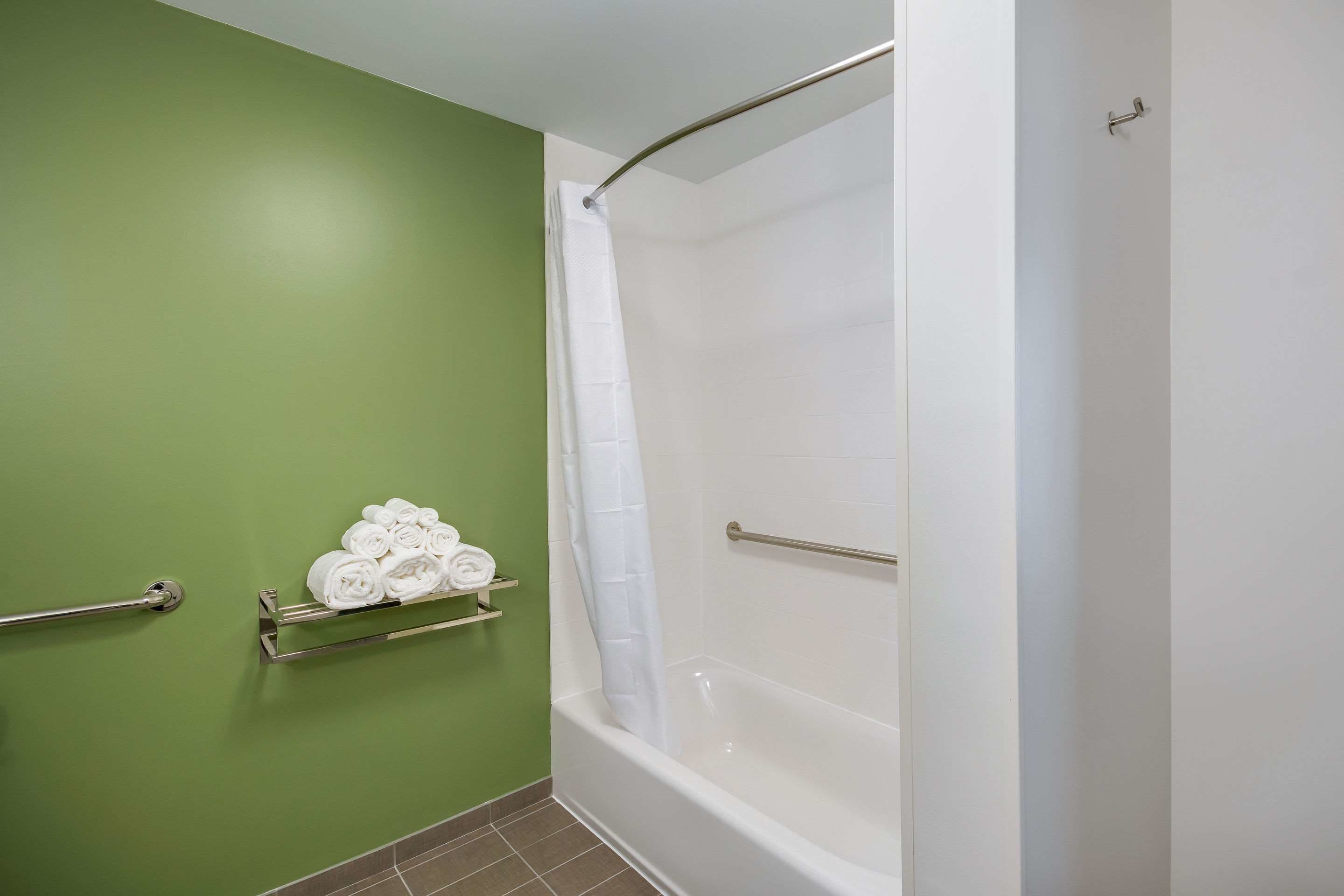 Un baño con pared verde, bañera blanca y cortina de ducha. Toallas ordenadas en un estante.