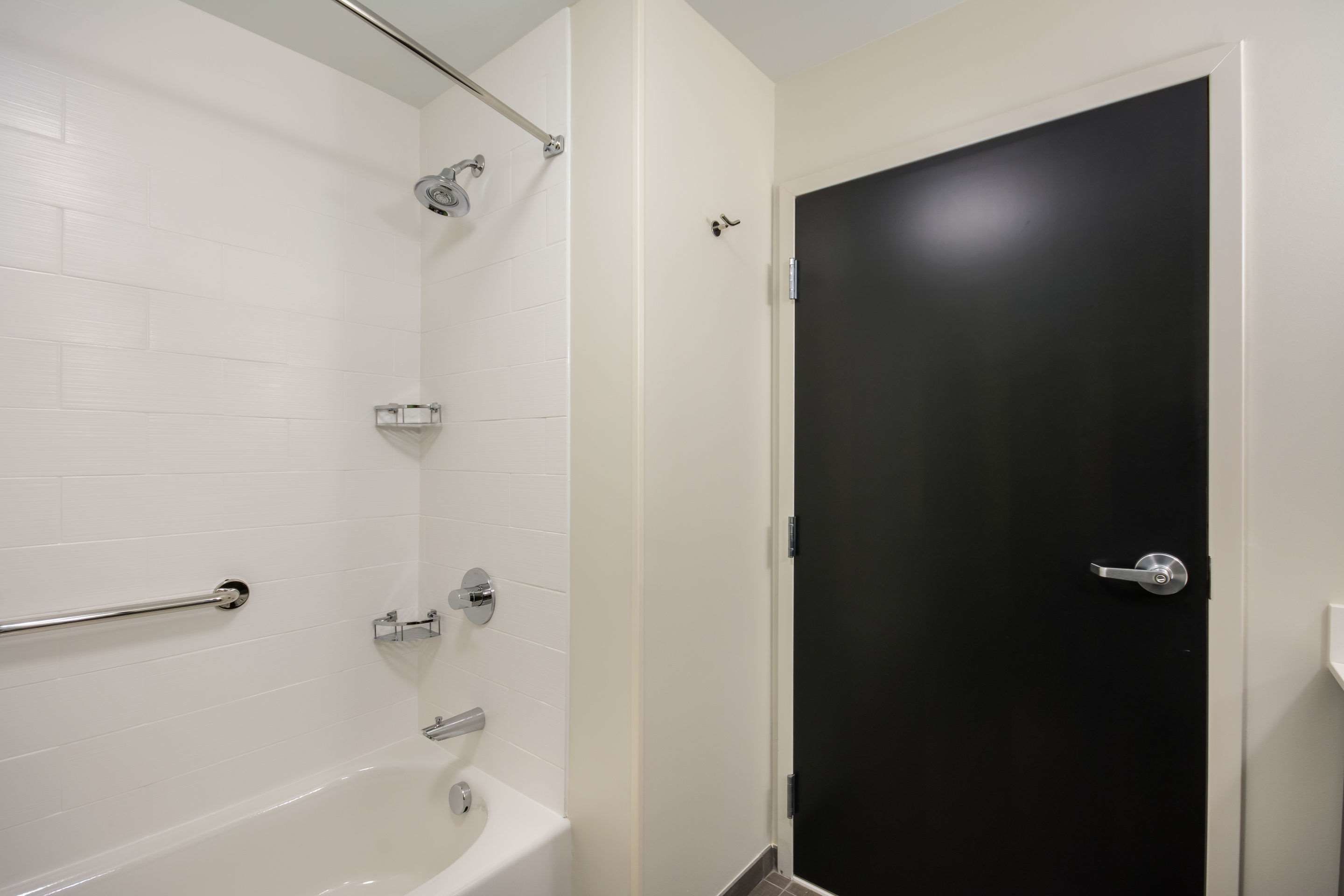 A bathroom with white tiled walls, a black door, a bathtub, and a shower area.