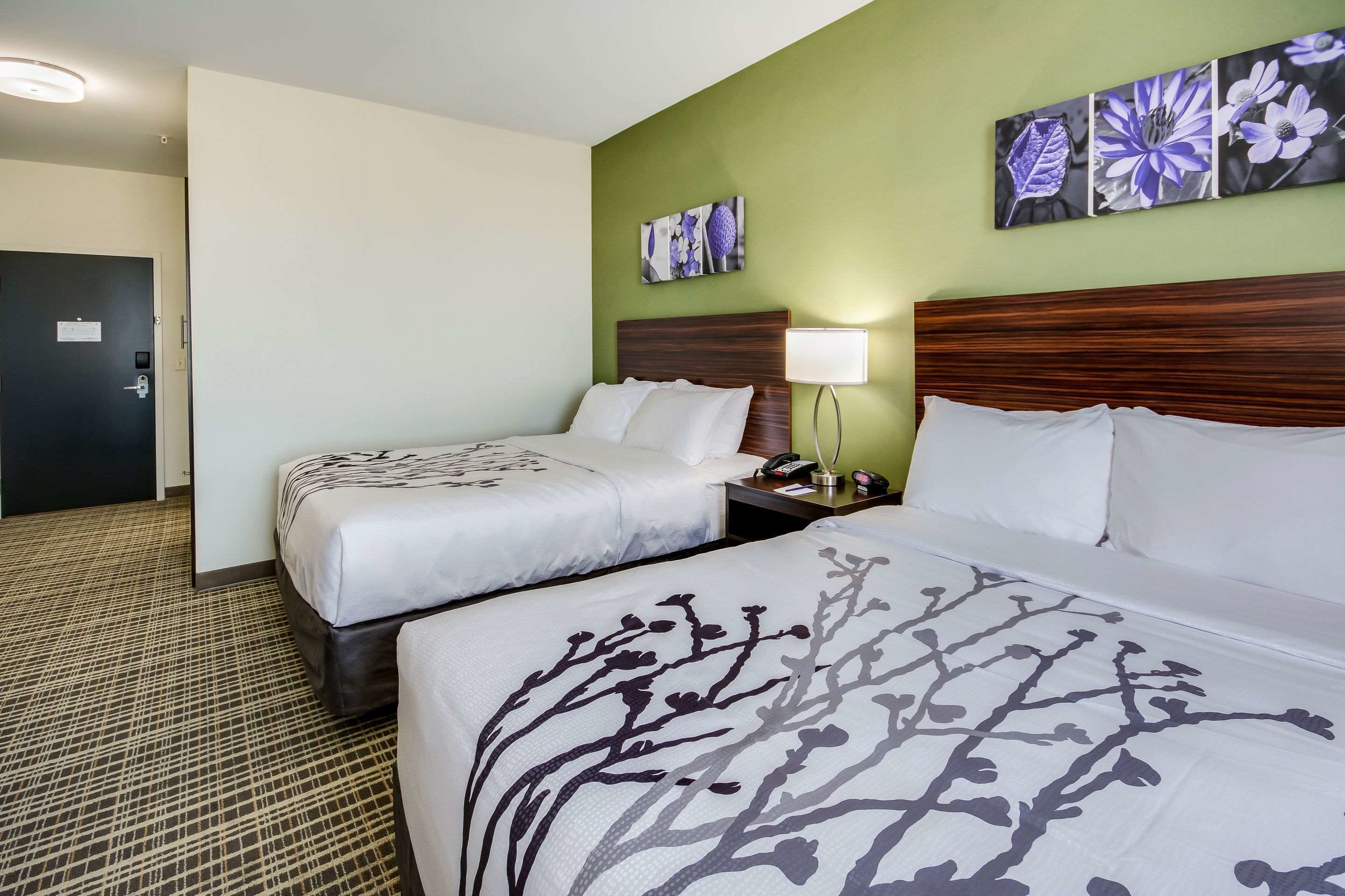 Habitación de hotel con dos camas, una mesita de noche, una lámpara y decoración floral en la pared.