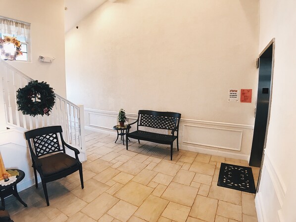 A hallway with two black chairs, a small table with a potted plant, a decorative wreath on the wall, and a staircase with a white railing. - Panoramic Ocean Views, 2 Large Decks, and Direct Beach Access! (North Wildwood)