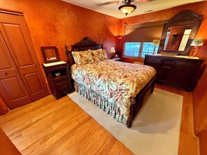Un dormitorio con piso de madera, una cama con colcha estampada, una mesita de noche, un aparador, un espejo y un ventilador de techo.