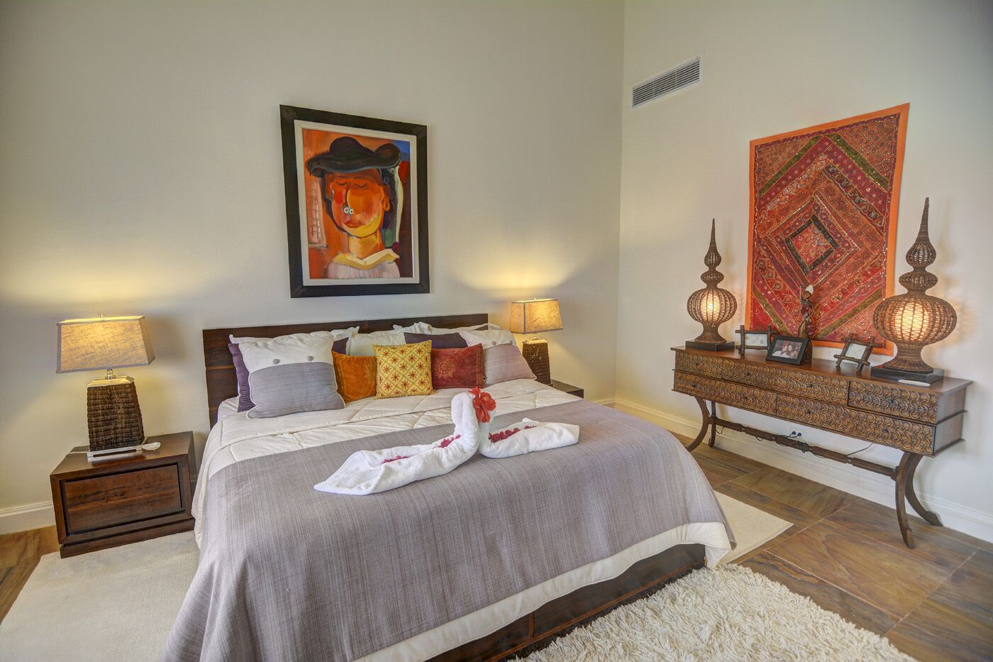 A neatly arranged bedroom with a bed, two bedside tables, a dresser, and a wall with a painting and a tapestry.