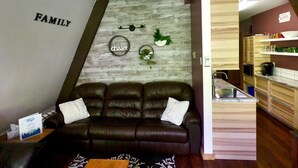 Een woonkamer met een donkerbruine leren bank, een klein tafeltje met een bord en een keuken met houten kasten en een aanrecht.