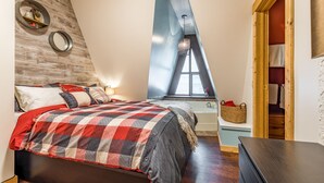 Een slaapkamer met schuine wanden, een bed met een geruite plaid, een houten hoofdbord, een klein raam met uitzicht en een houten garderobe.