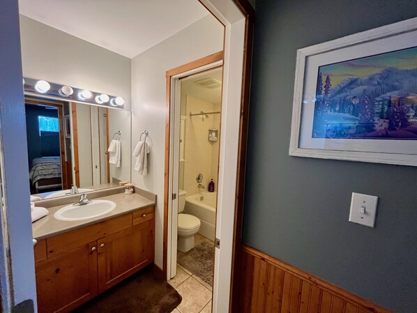 A bathroom with two sinks, a mirror with lights, a shower, and a painting on the wall.