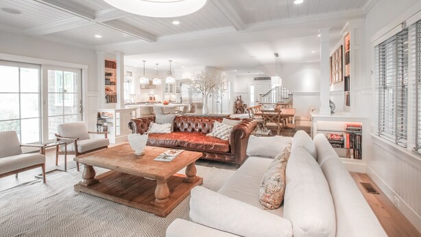 A spacious living room with a large brown leather sofa, a white sectional sofa, and a wooden coffee table. There is a kitchen area in the background with white cabinetry and a dining table. - 5 star, grand 8 BR estate, large yard, pool, pond, walk to town & beach (Nantucket)