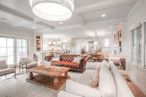 Een ruime woonkamer met een grote bruine leren bank, een witte hoekbank en een houten salontafel. Achtergrond toont een keuken met witte kastjes en een eettafel.