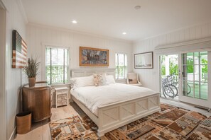 A bedroom with a large bed, a wooden cabinet, a small table, and a chair. There are windows with blinds and a view of bicycles outside.