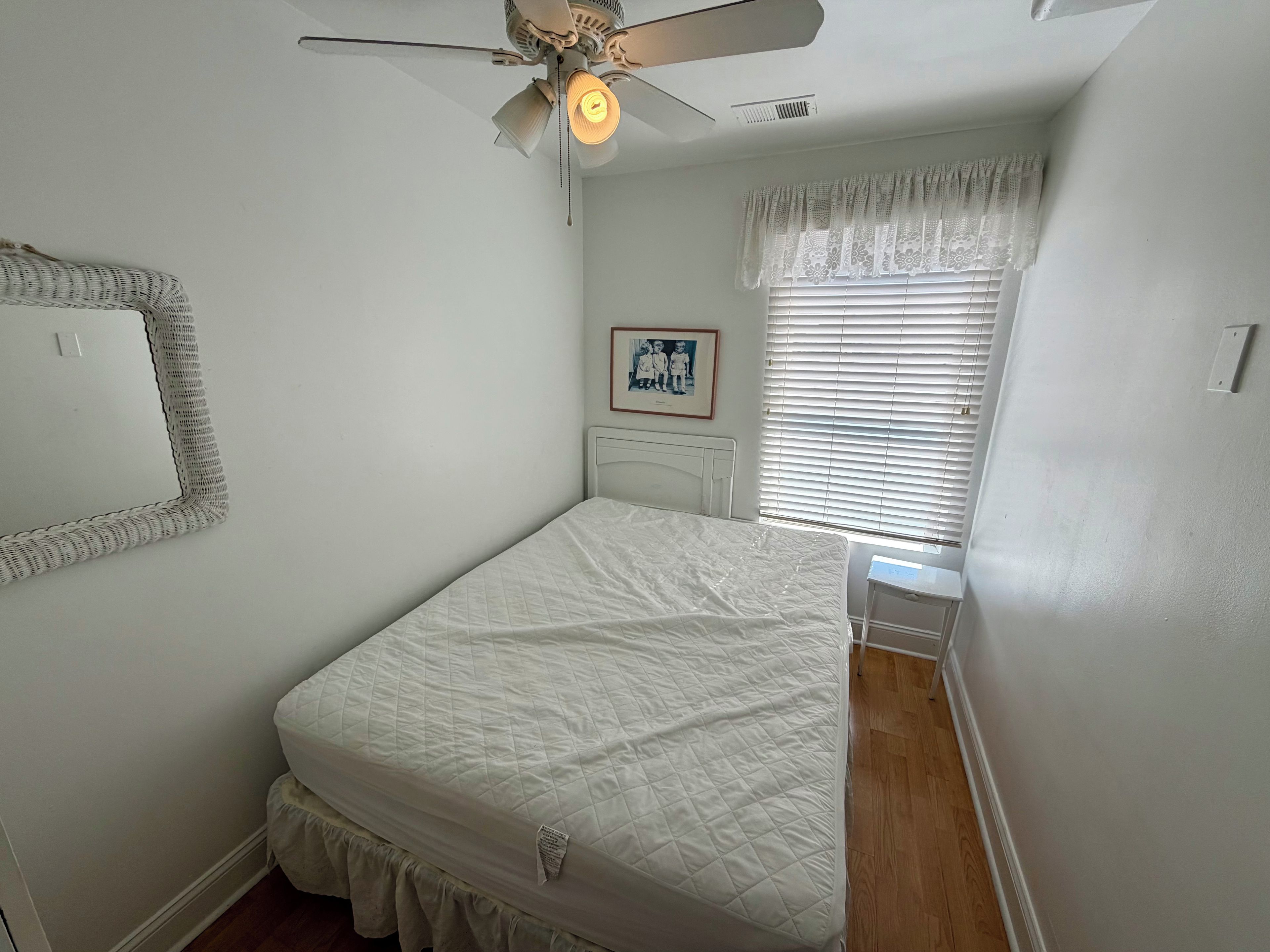 A bedroom with a bed, a ceiling fan, a window with blinds, a framed picture on the wall, and a bedside table.