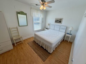 Ein Schlafzimmer mit einem Bett, einem Sessel, einem Nachttisch, einer Lampe, einem Deckenventilator und einem Fenster mit Jalousien.