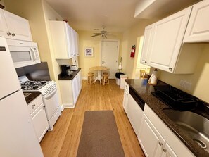 A compact kitchen with white cabinetry, a gas stove, a microwave, and a sink. There is a wooden dining table with stools and a ceiling fan.