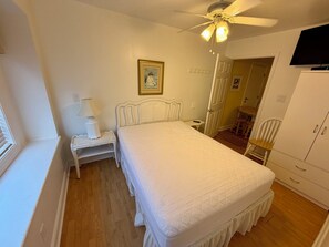 Quarto com cama, mesa de cabeceira, luminária, ventilador de teto, guarda-roupa e porta que dá para outro cômodo.