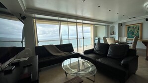 Ein Wohnzimmer mit einem Glastisch, zwei Sofas, einem Fernseher und einem großen Fenster mit Meerblick.