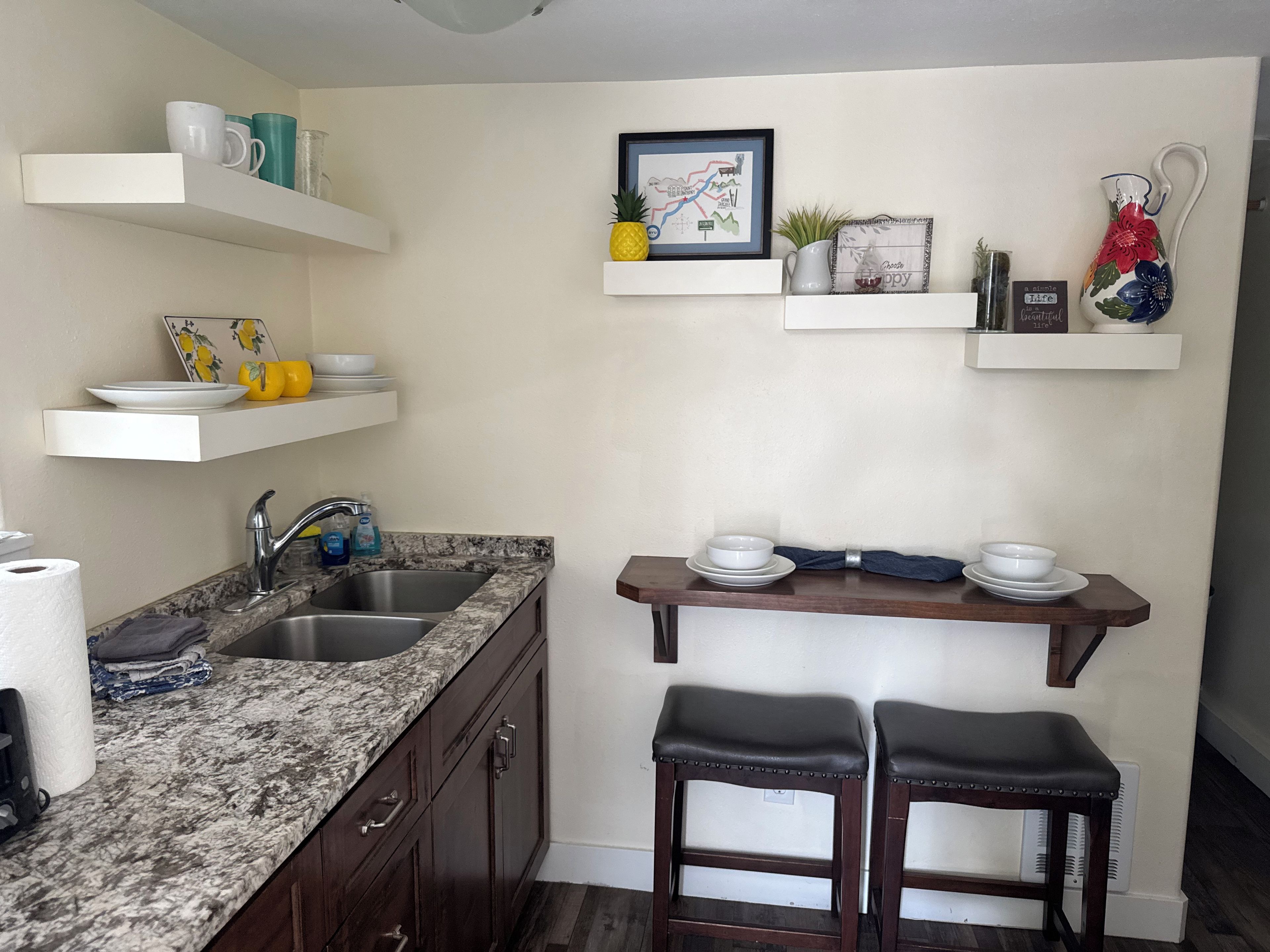 A kitchen with a granite countertop, stainless steel sink, and dark wooden cabinets. There are white shelves above the counter holding various kitchen items.