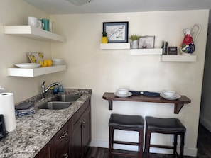 A kitchen with a granite countertop, stainless steel sink, and dark wooden cabinets. There are white shelves above the counter holding various kitchen items.