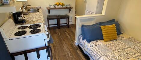 A compact kitchen with a stove, sink, and microwave, a small dining area with a table and chairs, and a bed with a patterned bedspread and pillows.