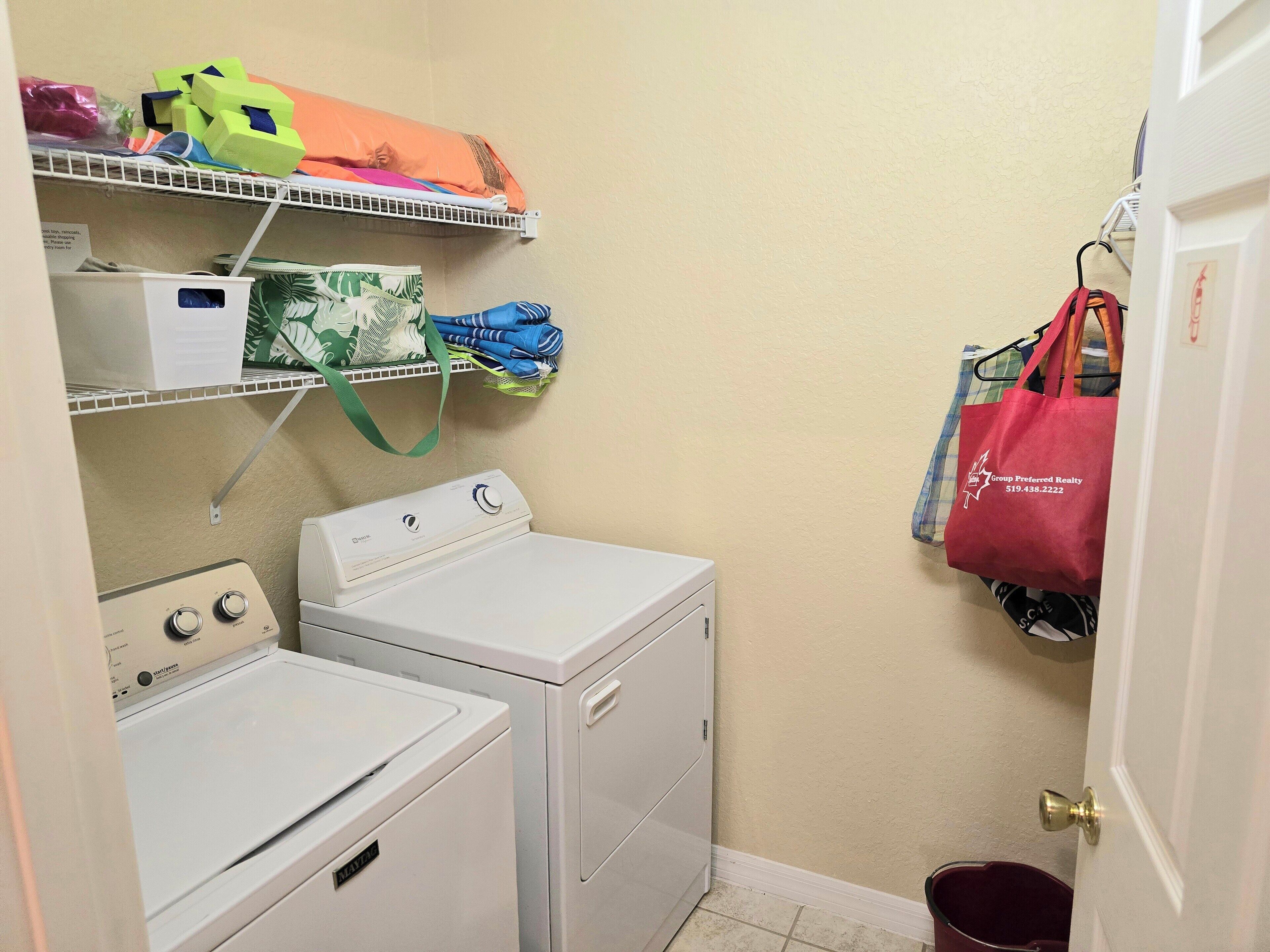 Un espace buanderie comprenant un lave-linge et un sèche-linge à chargement frontal, des étagères avec des vêtements pliés et des sacs, et un sac fourre-tout rouge accroché à un crochet.