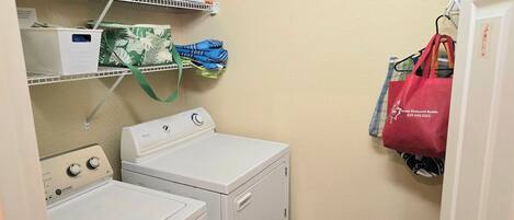 Un espace buanderie comprenant un lave-linge et un sèche-linge à chargement frontal, des étagères avec des vêtements pliés et des sacs, et un sac fourre-tout rouge accroché à un crochet.