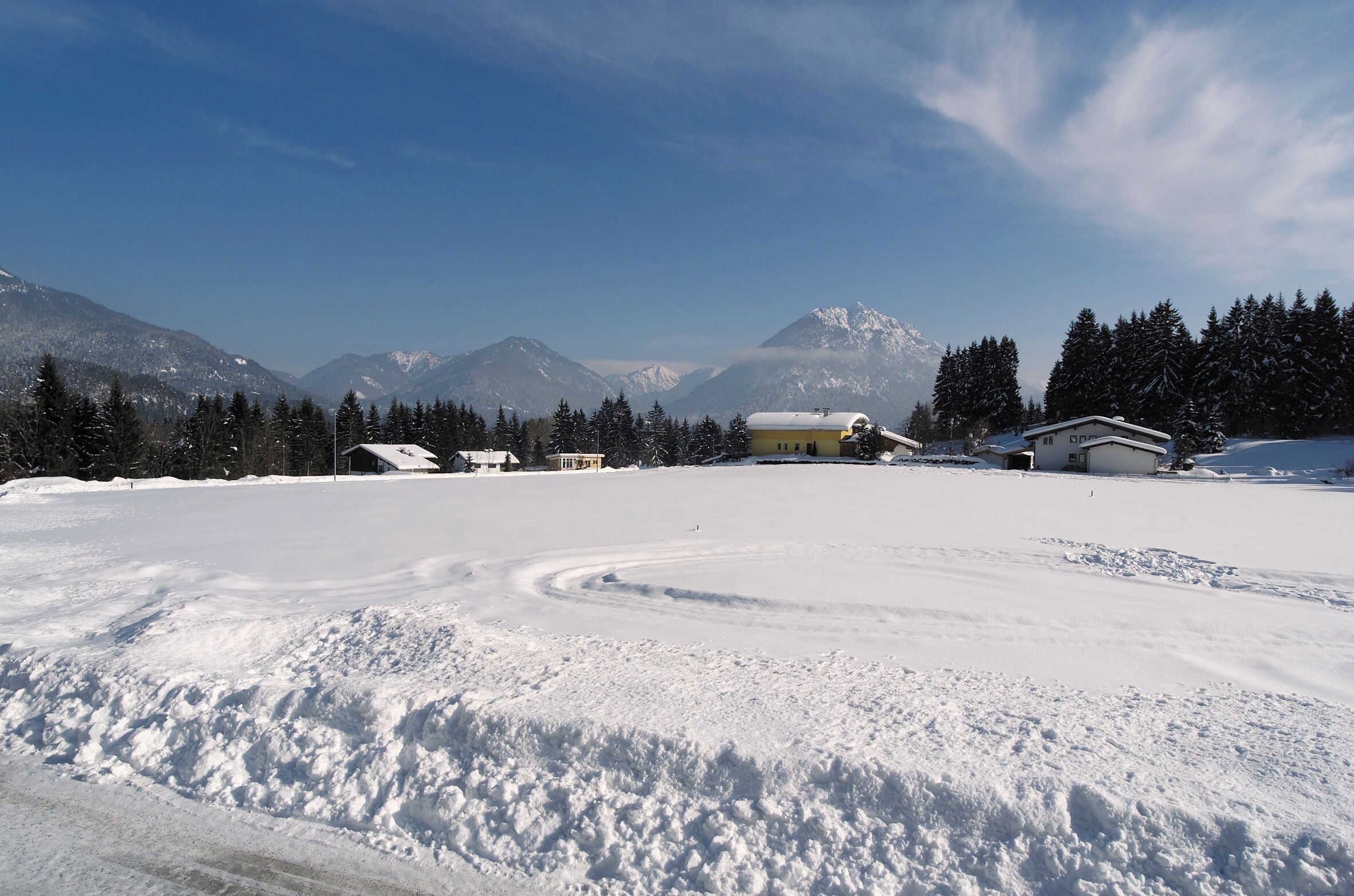 Paisagem coberta de neve com um edifício amarelo no centro, rodeado por montanhas e árvores.
