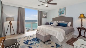 A bedroom with a bed, a ceiling fan, a view of the ocean, and a large window.