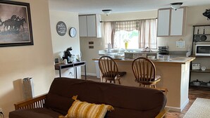 A cozy living room with a brown sofa, a coffee table, and a kitchen in the background. - The Farmhouse (Baton Rouge)