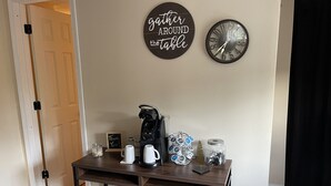 A room with a wooden table holding a coffee maker, cups, and a jar. A clock and a sign saying "Gather Around the Table" are on the wall. - The Farmhouse (Baton Rouge)