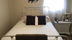 Una cama individual con estructura metálica, dos almohadas, una mesita de noche con lámpara, un jarrón pequeño con flores y una decoración de caballos en la pared.