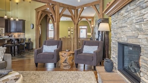 A spacious living room with a stone fireplace, wooden beams, and a dining area in the background.