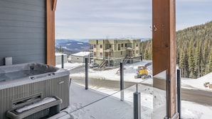 A hot tub on a balcony overlooking a snowy landscape with a building and a yellow vehicle in the background.
