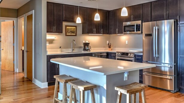 Una cocina moderna con armarios oscuros, electrodomésticos de acero inoxidable, iluminación colgante y una isla central con taburetes.