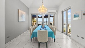 Een eetruimte met een lange tafel, stoelen, een kroonluchter en uitzicht op het strand door grote ramen.
