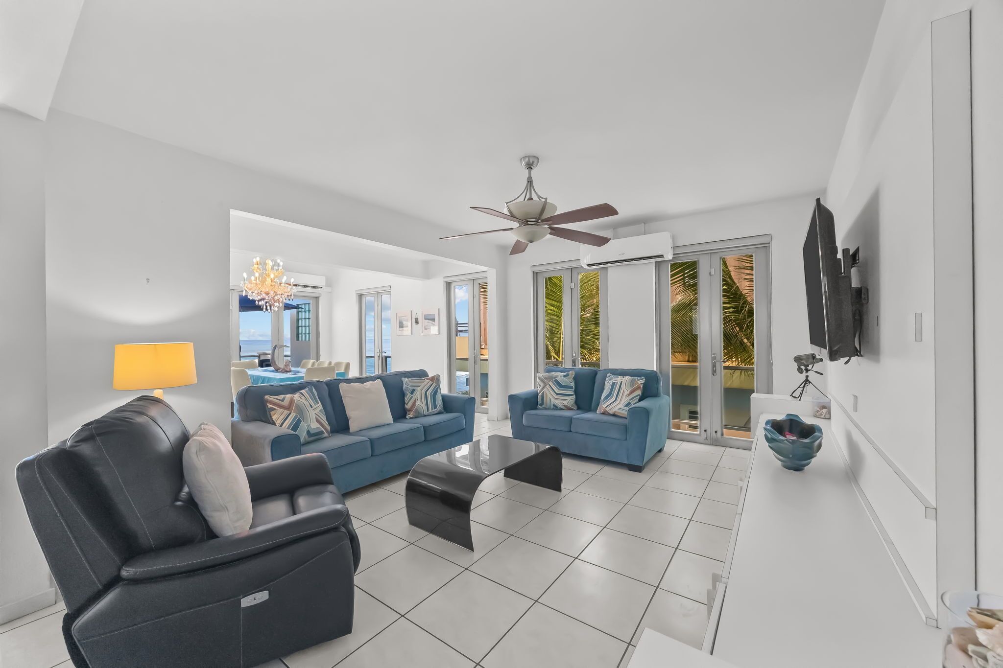 A modern living room with a black leather sofa, a blue sofa, a glass coffee table, and a ceiling fan.