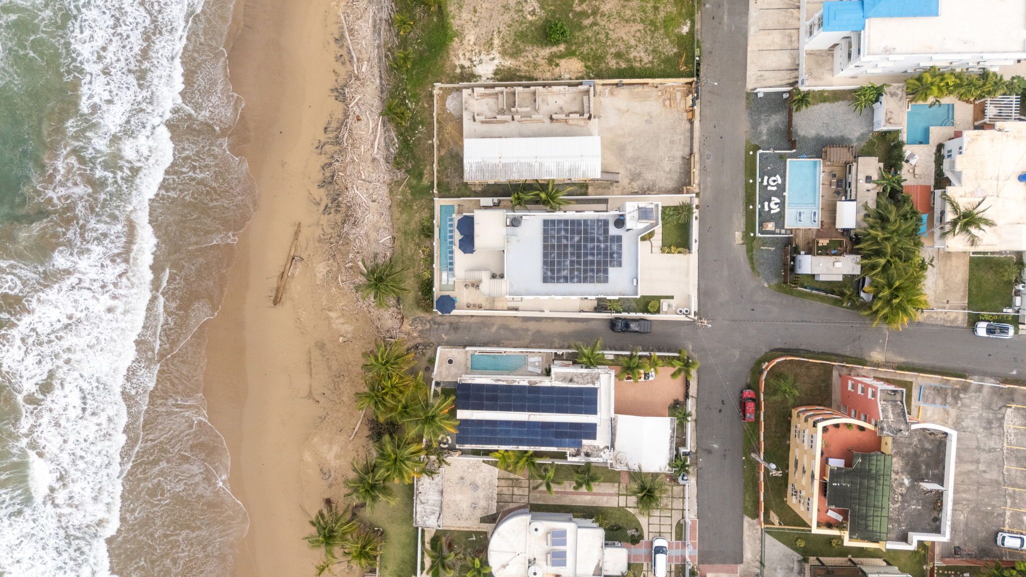 An aerial view of a coastal property with a beach, solar panels, and a swimming pool.