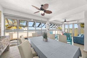 A spacious room with a large table, chairs, a ceiling fan, and large windows offering a view of palm trees.