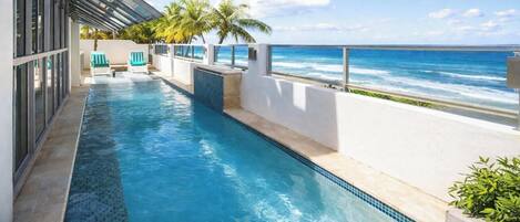 A swimming pool with a view of the ocean, a covered area with chairs, and a white wall.