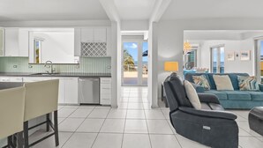 Een moderne keuken met witte kastjes, een eethoek met tafel en stoelen, een woonkamer met een bank en televisie, en een balkon met uitzicht op zee.