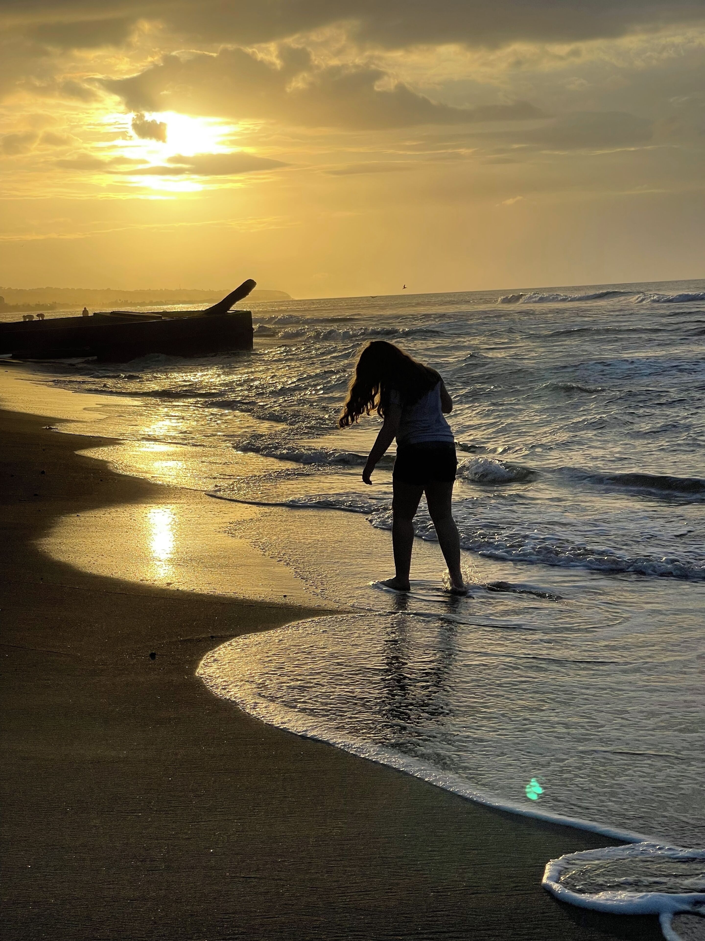 A beach scene with a person standing in the water, waves approaching the shore, and a sunset in the background.