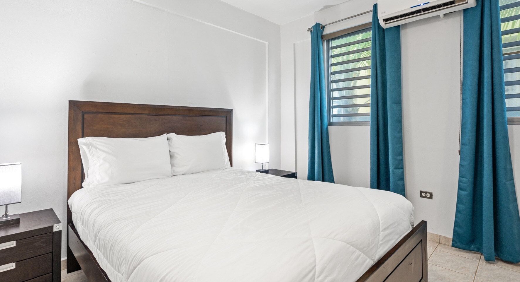 A neatly made bed with white bedding, a wooden headboard, and a nightstand with a lamp.