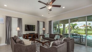 A spacious living room with a ceiling fan, a flat-screen TV, a fireplace, and a view of a swimming pool and palm trees.