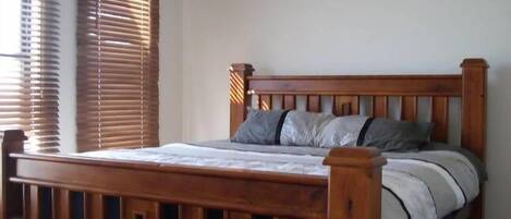 A wooden bed with a headboard, a window with blinds, and a plain wall.