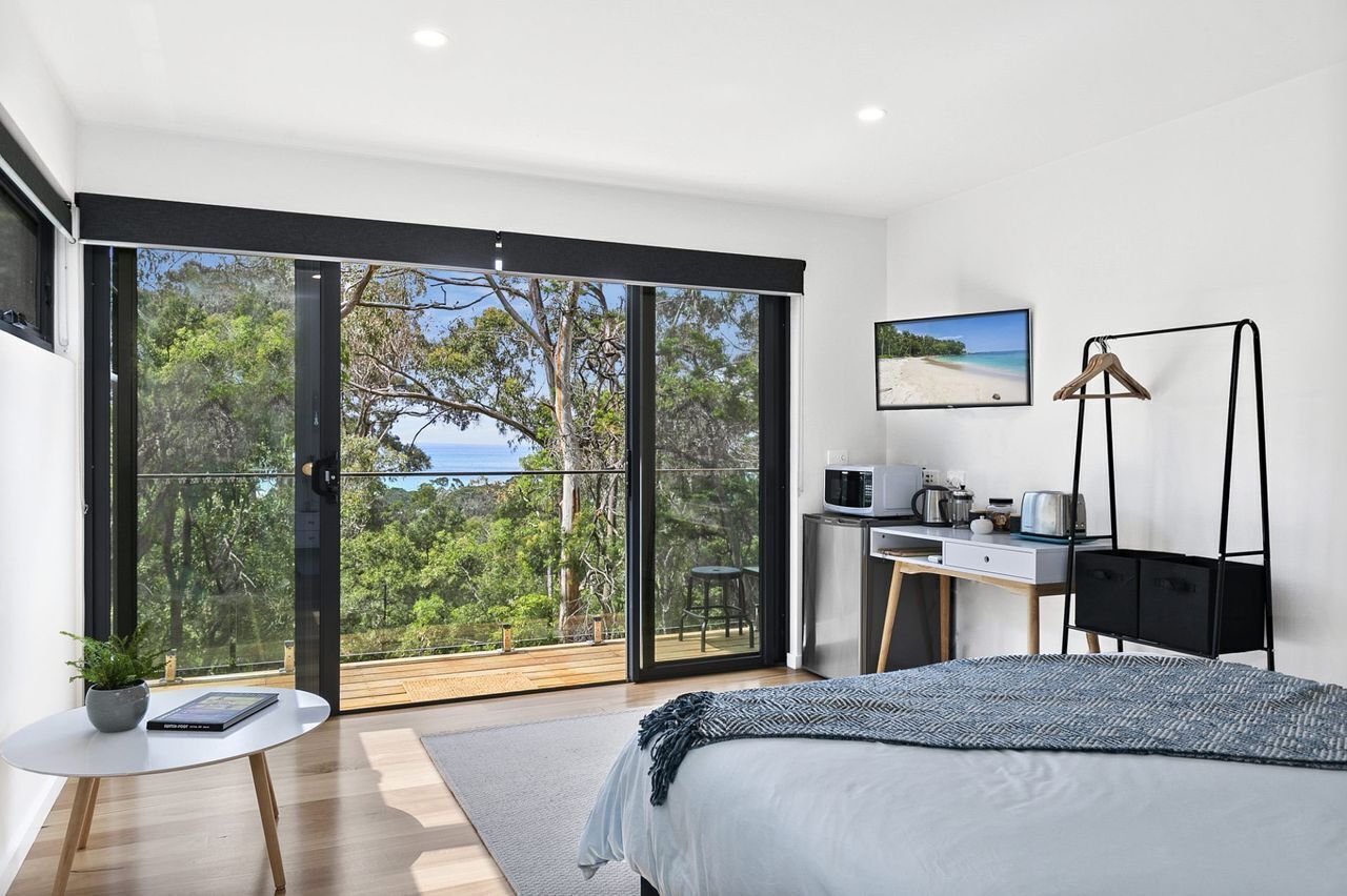A bedroom with a bed, a desk, a chair, a TV, and a balcony with a view of trees and the sky.