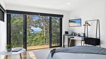 A bedroom with a bed, a desk, a chair, a TV, and a balcony with a view of trees and the sky.