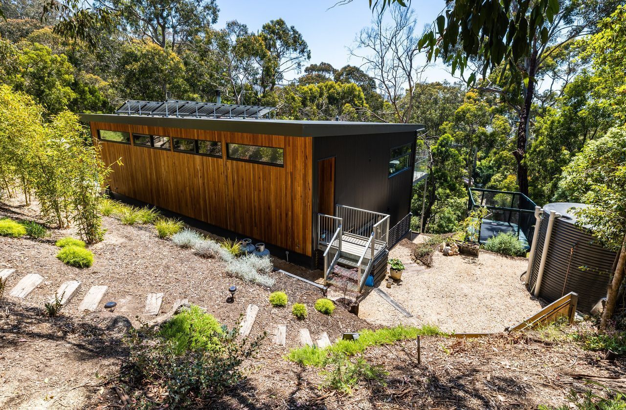 A modern building with a wooden exterior and a black roof, surrounded by a garden with various plants and a gravel path.