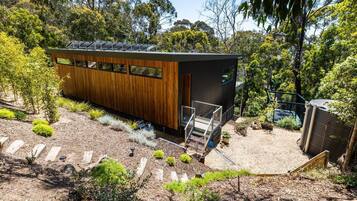 A modern building with a wooden exterior and a black roof, surrounded by a garden with various plants and a gravel path.