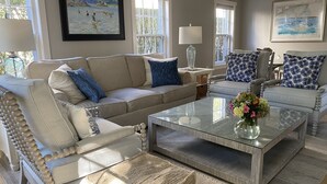 Een woonkamer met een beige bank, blauw-wit gestreepte kussens, een glazen salontafel met bloemen, en twee ingelijste schilderijen aan de wanden.