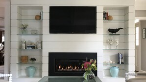 Een woonkamer met een open haard, een flatscreen-televisie erboven en glazen planken met diverse decoratieve objecten.