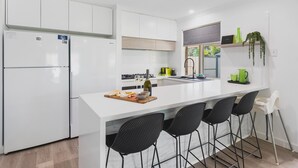 A modern kitchen with white cabinets, a central island with bar stools, a wine bottle, and a green plant. - Busselton Family Holiday House - by the bay (Busselton)