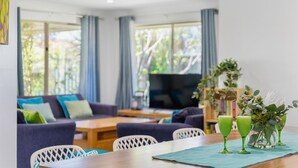 A modern living room with a sofa, dining table, and chairs. There is a TV, a plant, and a vase with flowers on the table. - Busselton Family Holiday House - by the bay (Busselton)