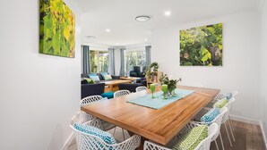A modern living room with a wooden dining table, white wicker chairs, a sofa, and a TV. - Busselton Family Holiday House - by the bay (Busselton)
