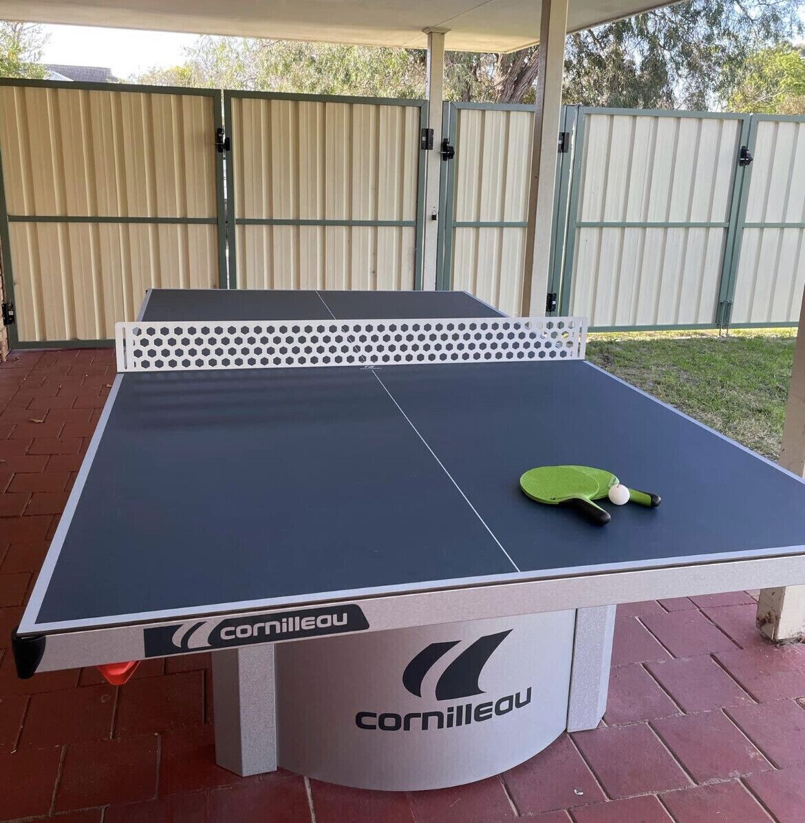 A Cornilleau ping pong table with a white net and a green paddle, set outdoors on a brick surface.